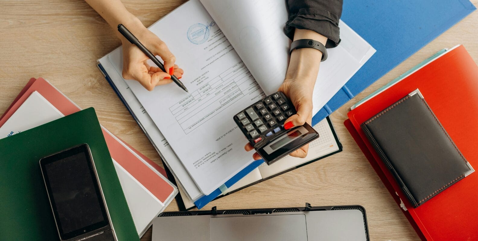 Prüfung durch das Finanzamt, Hände mit Taschenrechner und Ordner auf einem Tisch.
